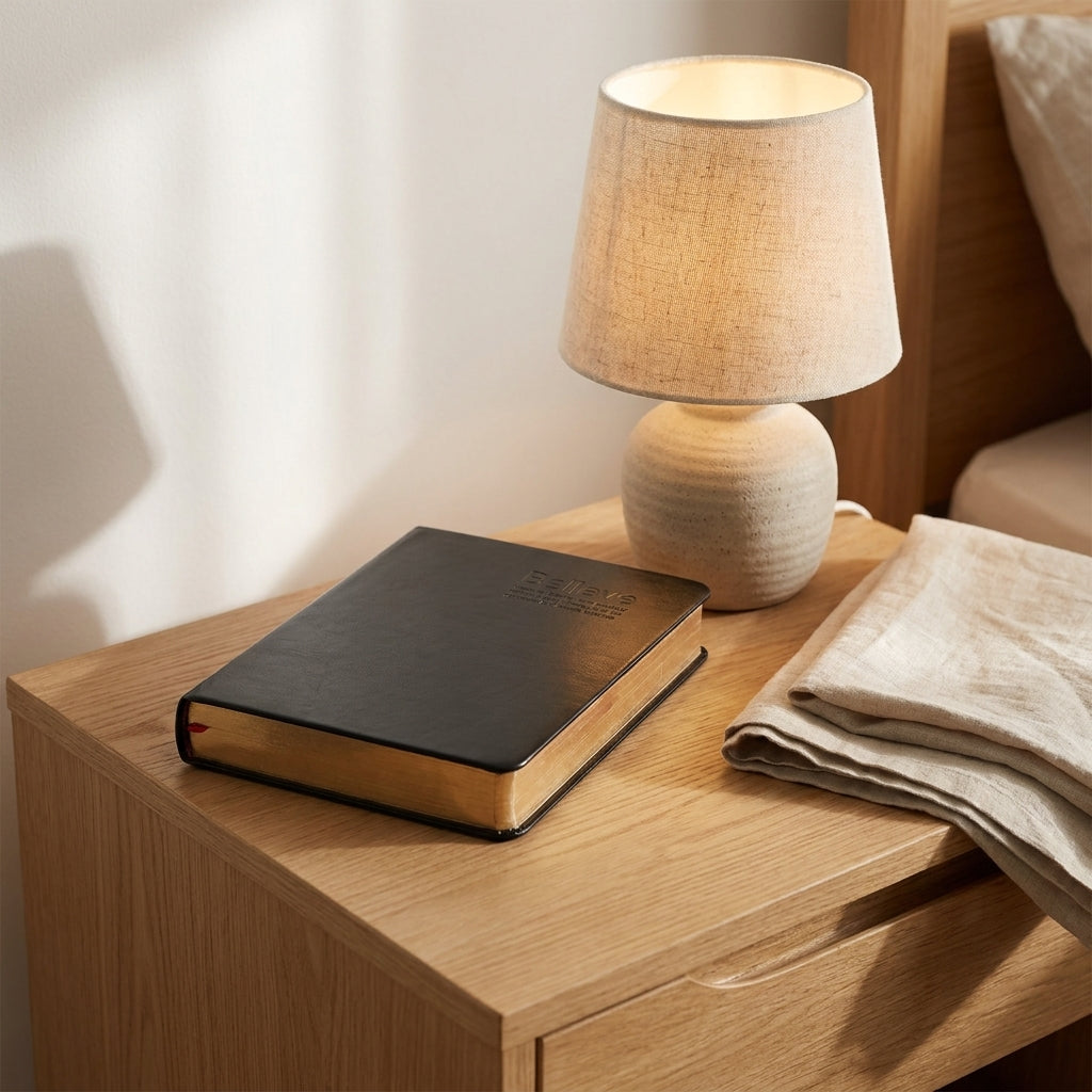 A lit table lamp with a textured beige fabric shade and matching base sits on a wooden bedside table next to a black hardcover book and a folded beige linen cloth.