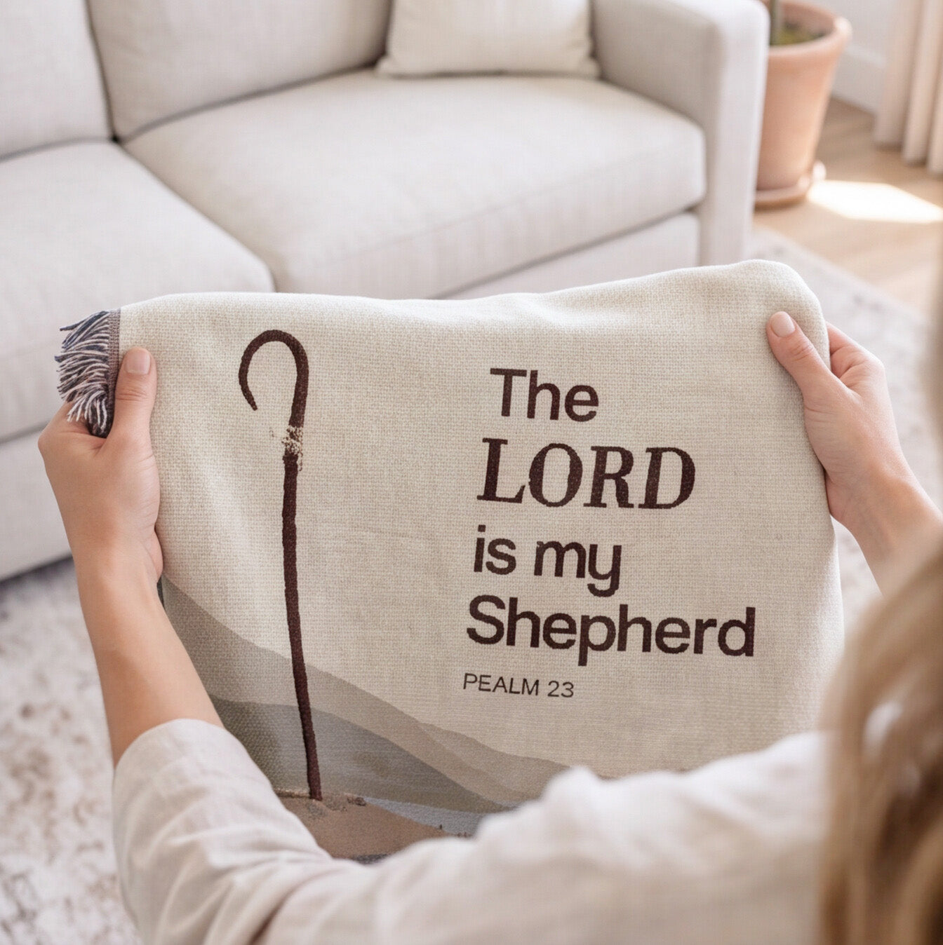 A person holds a cream-colored woven throw blanket featuring the text 'The LORD is my Shepherd' and 'PSALM 23' in dark brown lettering, alongside a shepherd staff graphic. The background shows a cozy living room setting with a light beige sofa.