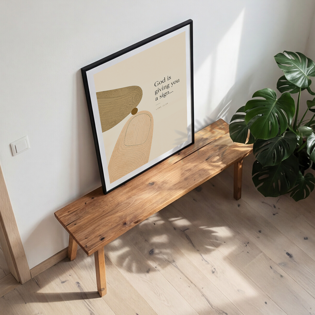 A framed abstract art print leaning on a rustic wooden bench in a bright room with a potted monstera plant nearby.