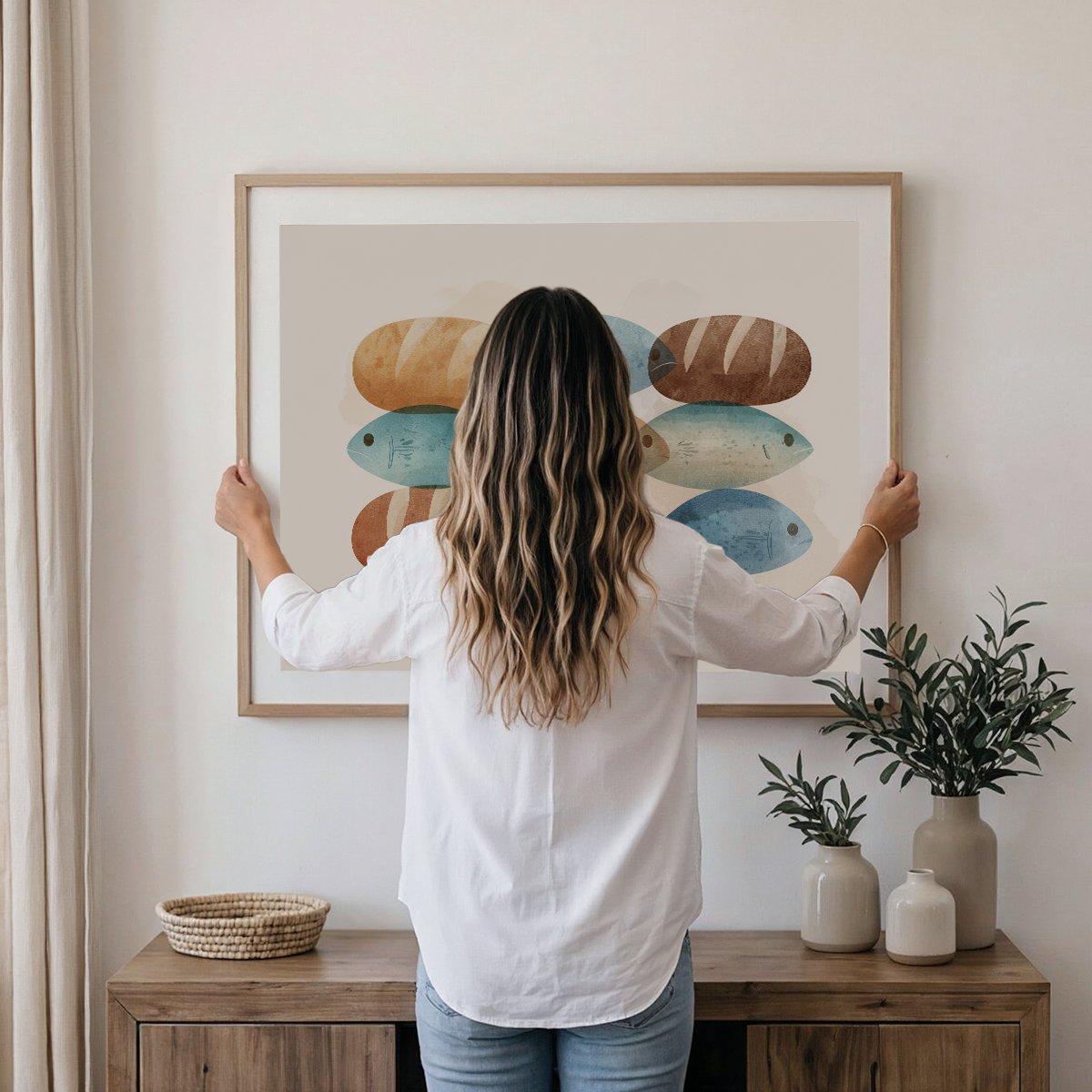 A woman with long highlighted hair stands with her back to the camera, holding a large framed art print on a white wall above a wooden console table. The print features an abstract watercolor design of fish in shades of blue, beige, and brown.