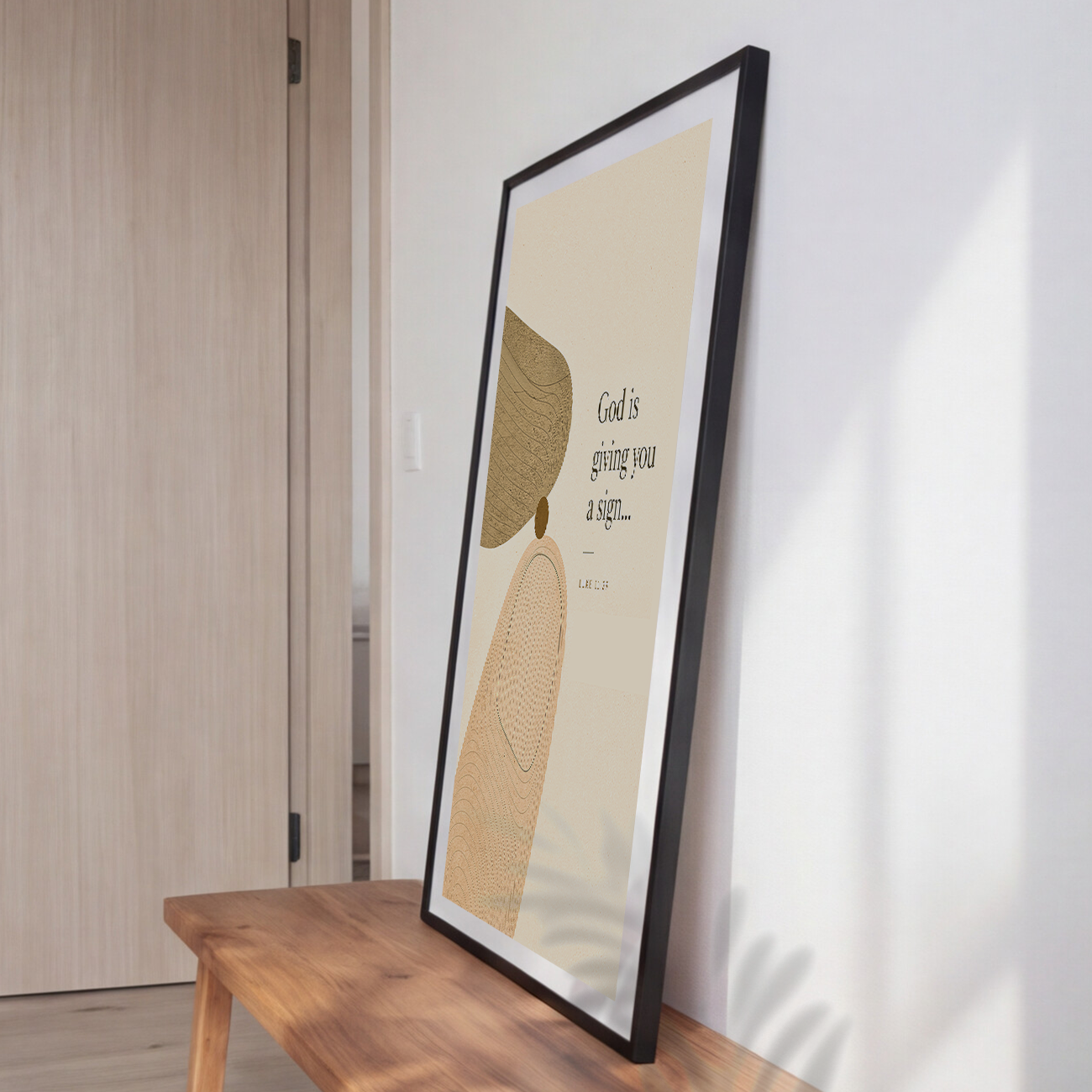A framed art print leaning against a white wall on a wooden bench in a modern room. The print features an abstract design with a beige background, a textured olive-green shape, a beige circular motif, and the quote 'God is giving you a sign.