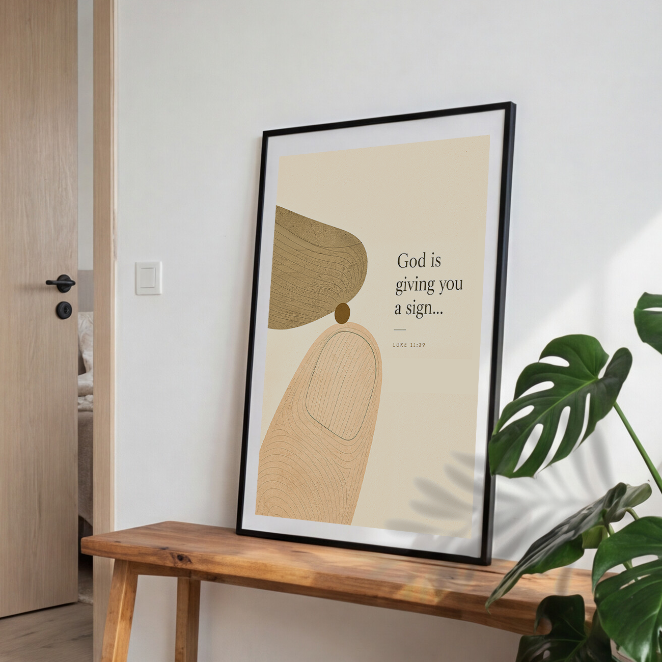 A framed minimalist art print leaning on a wooden bench against a white wall, with a green plant leaf in the foreground and a light wood door on the left. The print features abstract shapes and the text 'God is giving you a sign.