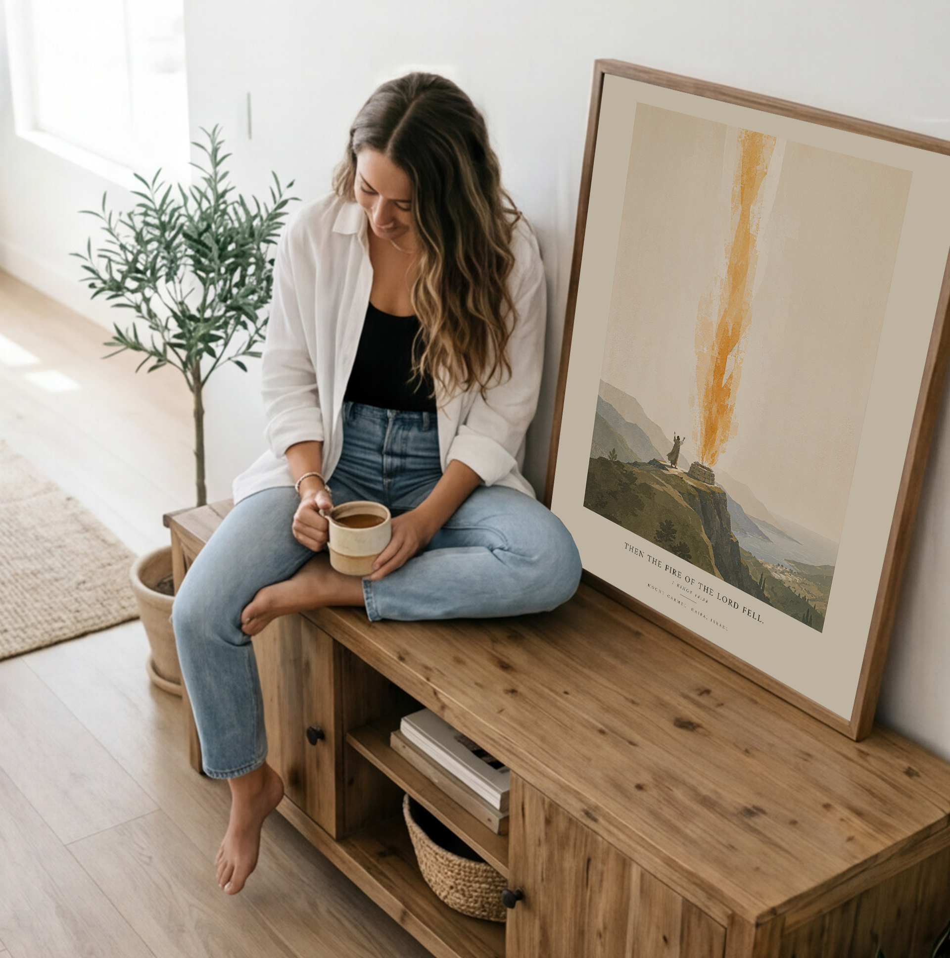 Woman sitting on a wooden cabinet holding a cup, with a plant and framed artwork in the background.