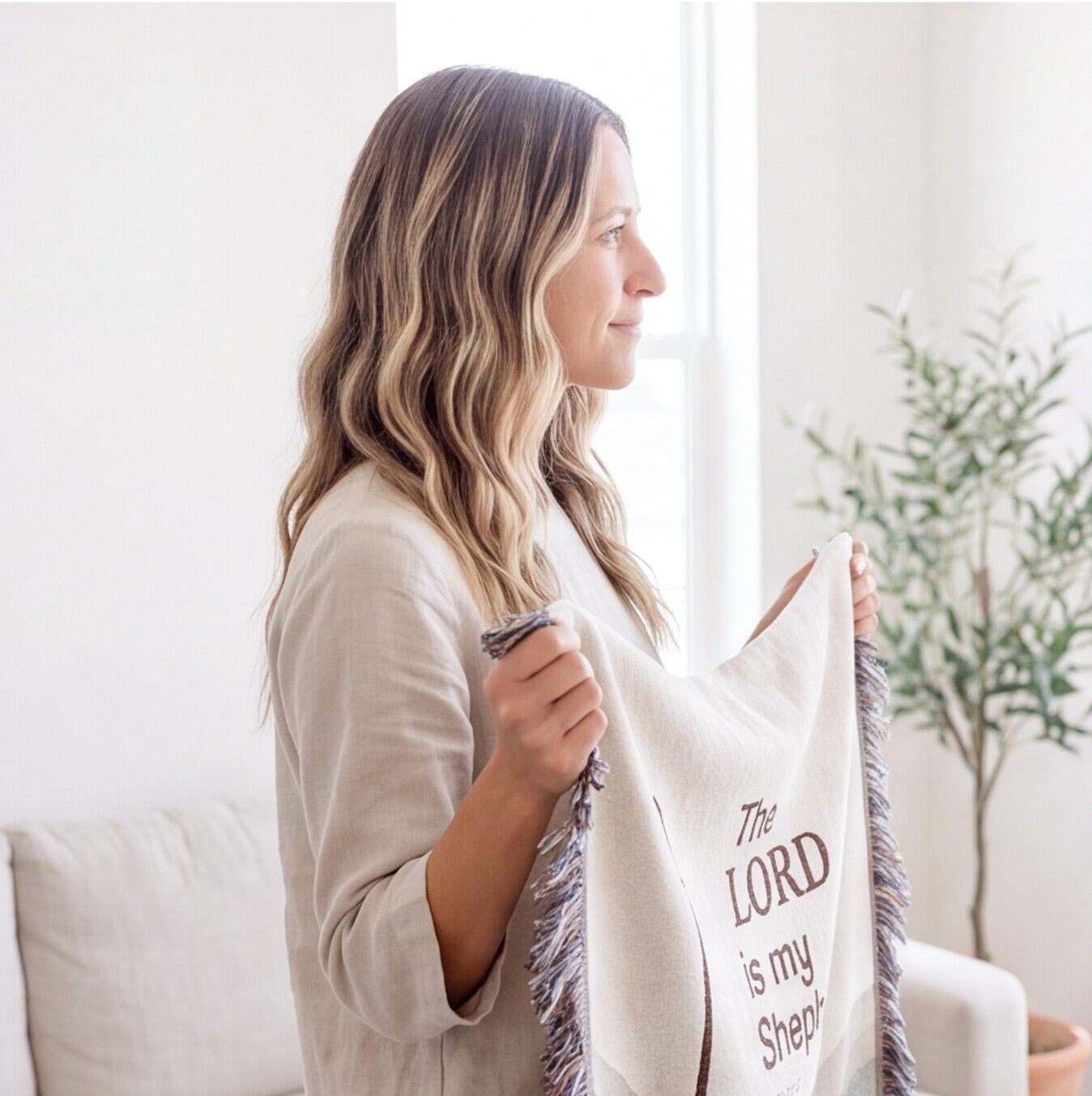 A woman in a beige top holds up a cream-colored throw blanket with fringe and the printed text 'The LORD is my Shepherd' in brown lettering, set in a bright, modern living room.