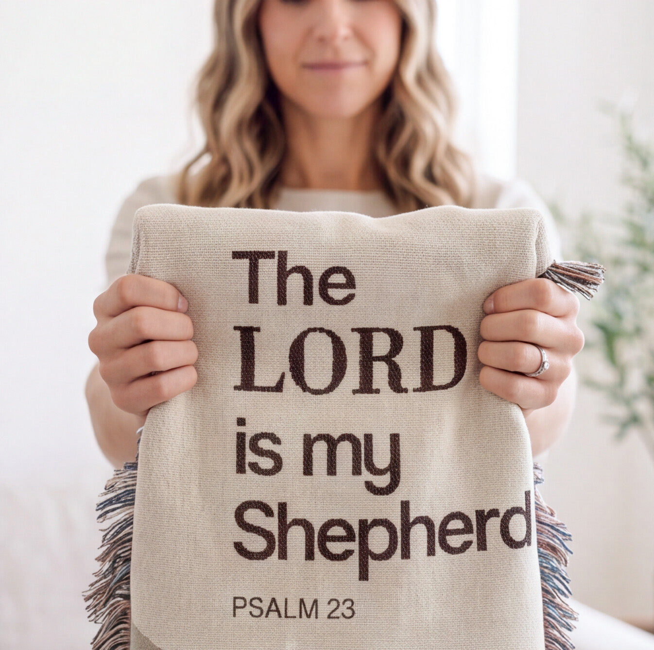A woman holds a beige linen throw blanket with dark brown text reading 'The LORD is my Shepherd PSALM 23'. The blanket has fringed edges and is shown in a bright, softly blurred indoor setting.