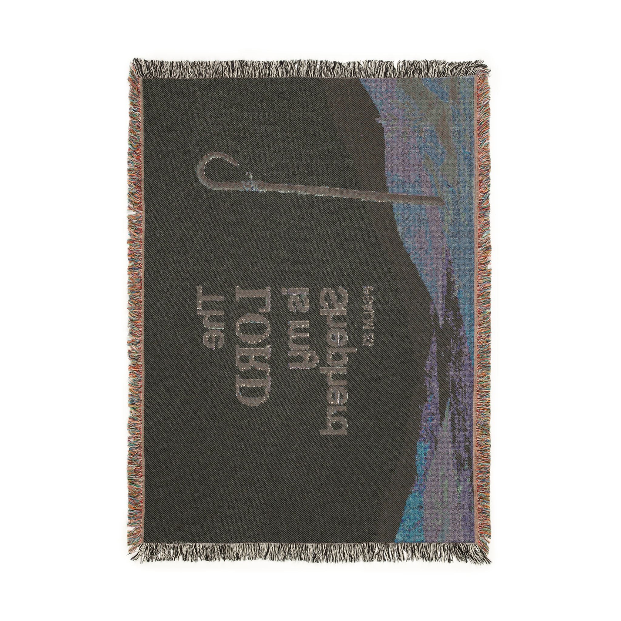 A rectangular woven throw blanket with fringed edges, featuring a dark charcoal background with a silver-toned sword hilt, the text 'The LORD Shema' from Exodus 34:7, and a blue-toned landscape illustration on the right side.