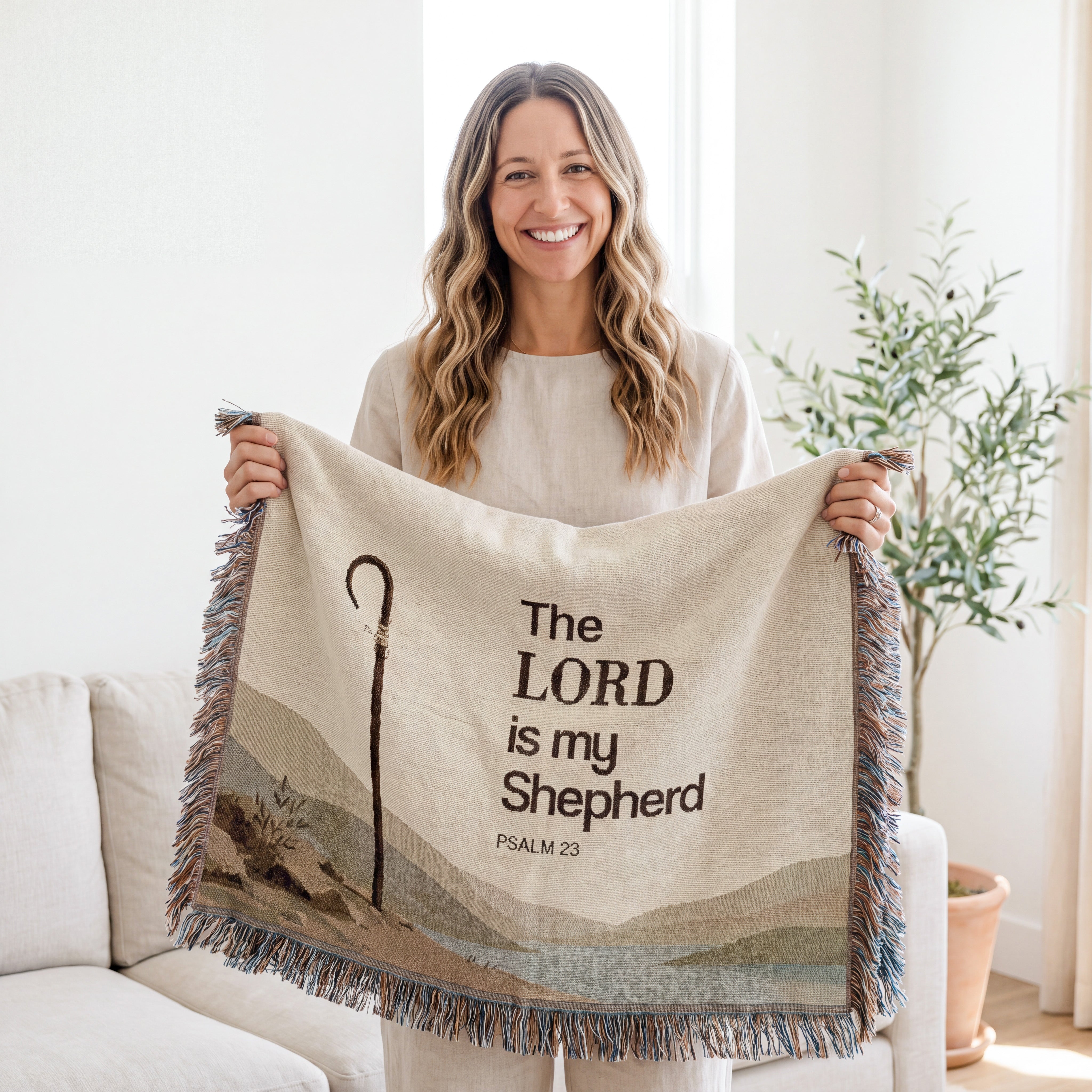 A smiling woman holds up a cream-colored woven throw blanket with fringe. The blanket features a printed design with a shepherd's staff, layered hills, and the text 'The LORD is my Shepherd Psalm 23'.