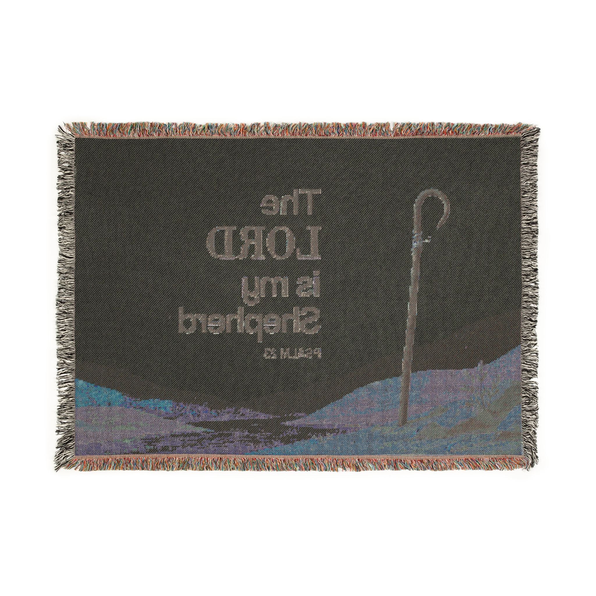 A rectangular woven throw blanket with fringed edges, featuring a dark charcoal background with the text 'The LORD is my Shepherd' from Psalm 23, alongside an illustration of a shepherd's staff and a blue-toned landscape.